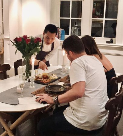 Server in an apron presenting a charcuterie board to a seated couple at a cozy candlelit home dining table, vase of red roses and plates visible.