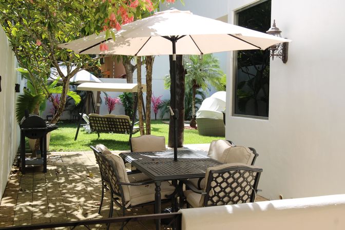 Sunlit backyard patio with a metal outdoor dining table and cushioned chairs under a large beige umbrella, a charcoal grill on pavers, green lawn with lounge chairs and tropical plants in a private garden courtyard.
