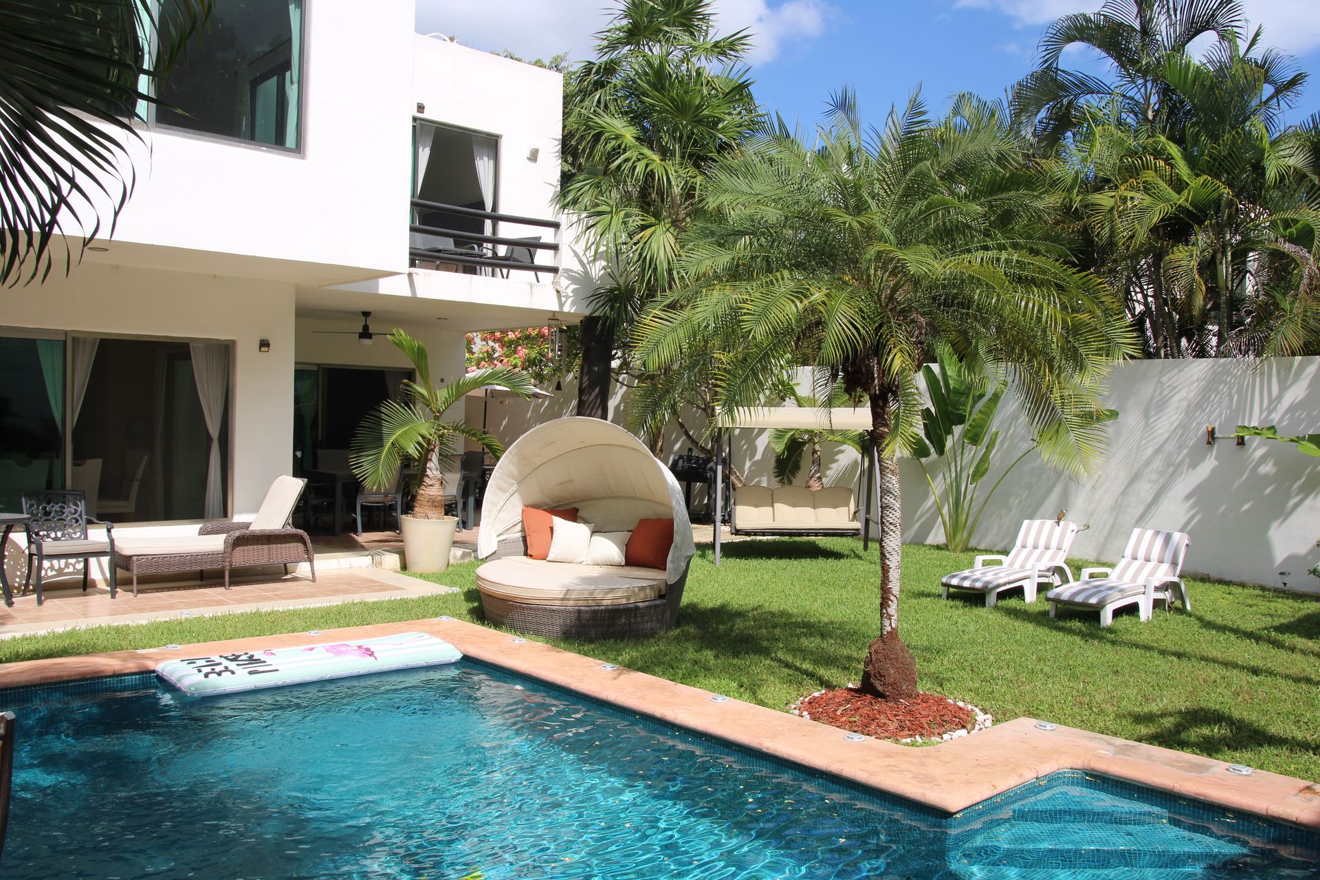 Sunny tropical villa backyard with turquoise swimming pool, round canopy daybed with orange pillows, palm trees, green lawn and striped lounge chairs.