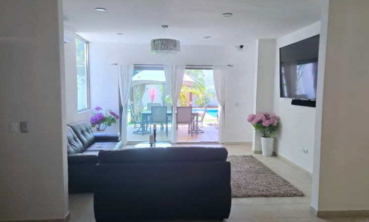 Bright modern living room with black leather sofa and wall-mounted TV, crystal chandelier, sliding glass doors to a sunlit patio dining area and backyard pool, decorative pink potted flowers and shag rug.