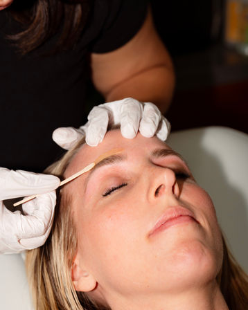 Close-up of a relaxed client receiving eyebrow waxing at a beauty salon — technician in white gloves applies warm wax with a wooden spatula to shape brows