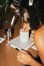 Close-up of hands setting a wick into a white ribbed candle jar on a tray at a DIY candle-making workspace, with a stainless pouring pitcher, thermometer and craft tools on a wooden table.