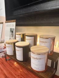 Artisan soy candles in glass jars with wooden lids arranged on a tiered wooden display, warm fireplace glow and orange fabric creating a cozy boutique market vibe.