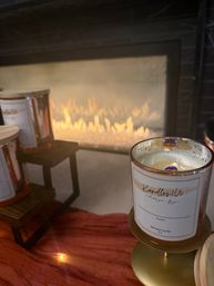 Lit soy-wax candle in a glass jar on a gold stand beside a modern gas fireplace, with extra candle jars on a wooden display and a rust-colored throw for a cozy living-room vibe.