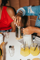 Close-up of hands stirring a metal pitcher over a mug at a café table, denim-sleeved person preparing a drink with two yellow citrus beverages with black straws, small glass bottles and a digital thermometer nearby.