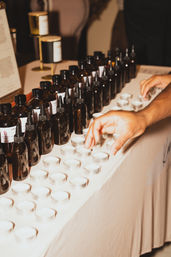 Hand reaching to pick a tealight tester at a fragrance sampling table lined with metal tealight cups and amber glass dropper bottles on a white tablecloth — artisanal candle and scent sampling station.