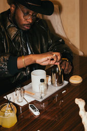 Person wearing a black cowboy hat and leather jacket stirring an artisanal cocktail on a polished wooden table with glass vials, a metal mixing cup, digital thermometer and a citrus drink — stylish lounge bartending scene