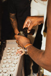 Close-up of hands pouring amber liquid from a brown bottle into a small glass vial beside rows of small sample tins, suggesting artisan fragrance or essential oil sampling.