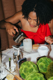 Person in a red top pours liquid from a stainless pitcher into a ribbed jar at a DIY candle-making table with a thermometer, wooden lid, takeaway coffee cup and green moss decor.