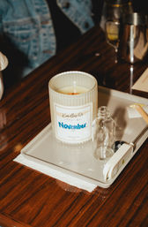 Ribbed white jar candle labeled "November" on a gray tray with a small clear glass bottle and electric lighter, arranged on a glossy wooden table — cozy tabletop candle display.