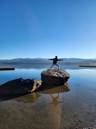 Person doing a yoga warrior pose with arms outstretched on a sunlit rock at a calm mountain lake, clear blue sky, distant mountains and crisp reflection in the water.