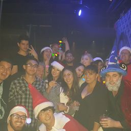 Lively holiday party in a dim nightclub with a group wearing Santa hats and festive costumes, colorful string lights, drinks in hand, and a man wearing a blue wizard hat.