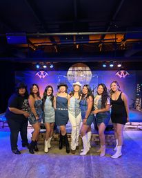 Eight women in denim and cowgirl boots smiling and posing on a lit stage at a country-music venue with purple-blue lighting and a rustic wooden backdrop — fun night out.