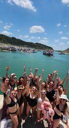 Sunny lake boat party: a group in swimsuits and heart-shaped sunglasses cheering with drinks on a pontoon, turquoise water, rocky shoreline and nearby party boats with slides.