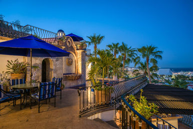 Oceanfront terrace at dusk with palm trees, wrought-iron railings, blue umbrellas and striped patio chairs, decorative stone fountain, overlooking coastal city lights and sea