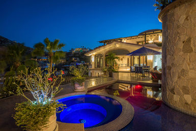 Luxury hillside villa terrace at dusk with a glowing blue spa and reflecting pool, outdoor dining under an umbrella, potted palms and stone-clad walls