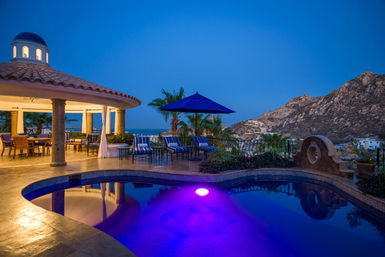 Oceanfront luxury villa pool at dusk with a purple-lit pool, Mediterranean-style gazebo, striped lounge chairs and rocky coastline view.