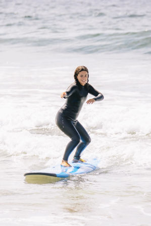 Smiling surfer in a black wetsuit balancing on a blue surfboard while riding a small wave along a sunny ocean beach