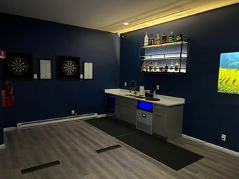 Modern home bar and game room with navy blue walls, illuminated floating liquor shelf, sink and mini-fridge, two dartboards and light wood flooring.