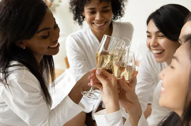 Four friends in white robes smiling and toasting with champagne flutes in a bright indoor bachelorette or bridal-party celebration.