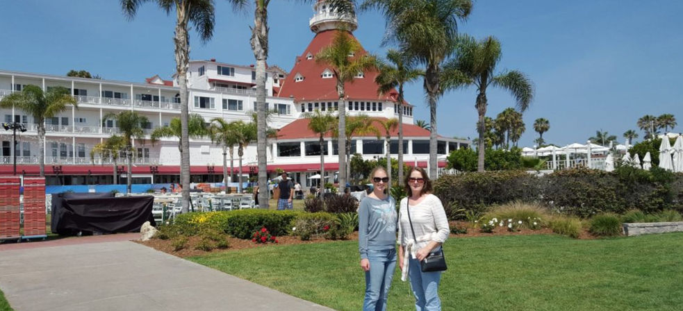 Two friends posing on a sunny lawn in front of a red-roofed Victorian seaside hotel with palm trees under a clear blue California sky.