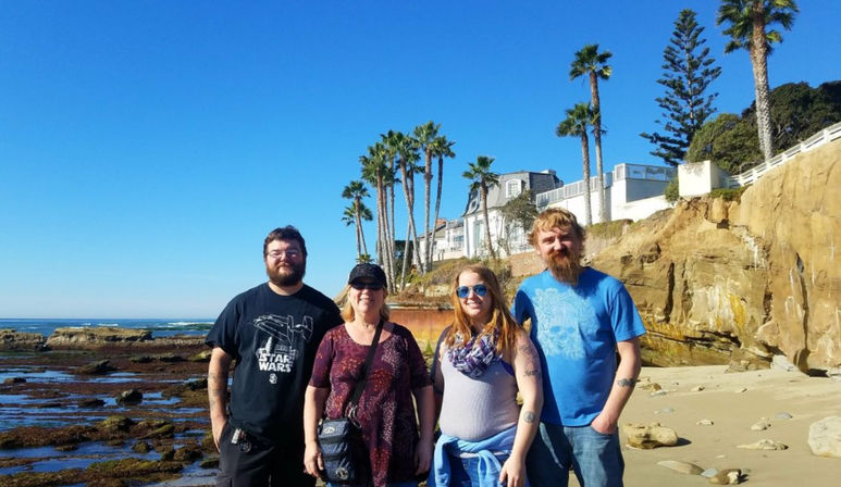 Smiling group of four adults posing on a sunny coastal beach with rocky tide pools, sandstone cliffs, tall palm trees and a bright blue ocean under a clear sky.