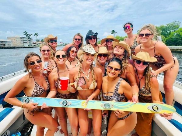 Group of women in animal-print bikinis and sun hats partying on a motorboat, smiling and holding a colorful wakeboard with built-in cup holders, palm-lined coastal waterway and cloudy sky in the background.