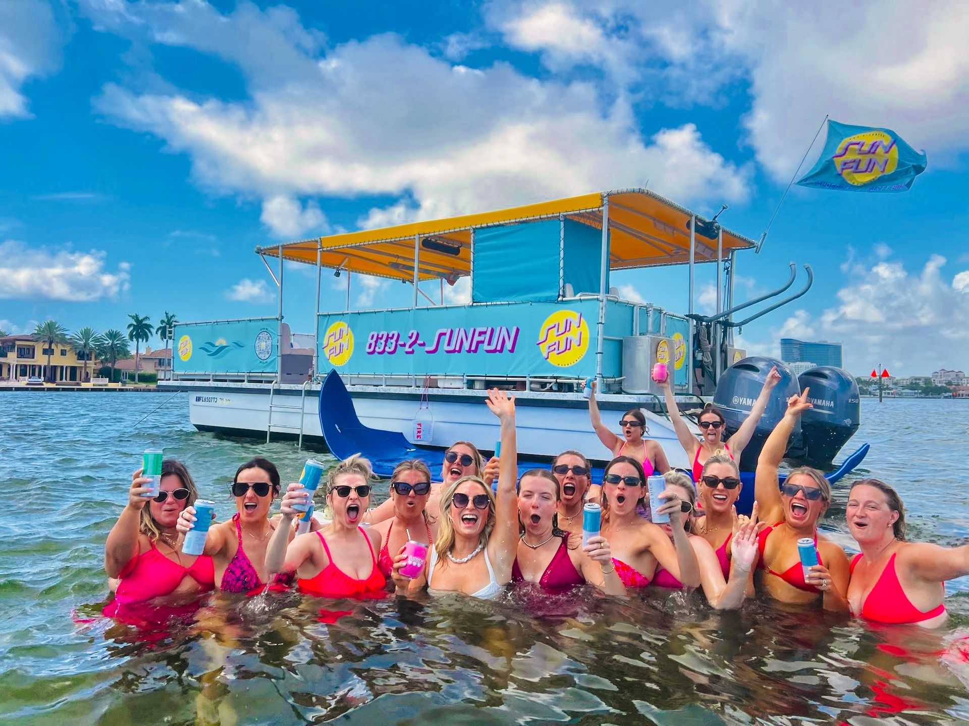 Cheerful group of women in colorful swimsuits waist-deep in a sunny coastal bay, holding canned drinks and cheering in front of a bright, decorated party pontoon boat with palm-lined shoreline