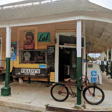 Yellow neighborhood corner store with covered porch, wooden bench, colorful hand-painted signs, chalkboard specials and an ice cooler, a red vintage bicycle locked to a green post in front.