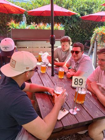 Group of friends enjoying large beer mugs at a wooden picnic table on a sunny outdoor patio with red umbrellas, planters, and table number 89.