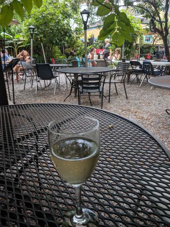 Cozy outdoor garden patio with metal mesh bistro tables on gravel, leafy canopy and diners in the background, white wine glass in the foreground.