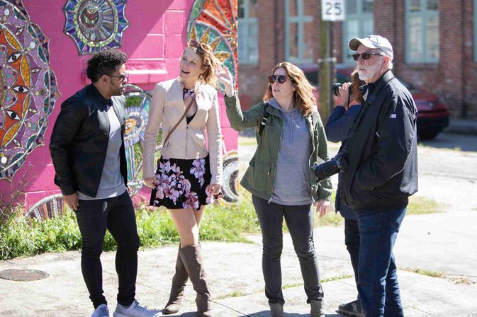 Four adults on a sunny urban sidewalk beside a colorful mosaic street-art mural on a pink wall; two women and two men chatting—one woman pointing, one man holding a camera.