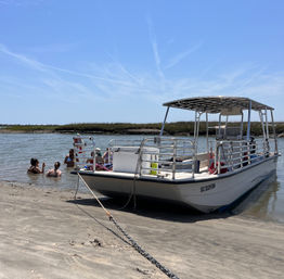 Pontoon boat beached on a sandy tidal inlet with people wading in shallow water, coastal marsh grasses and a clear blue sky on a sunny day