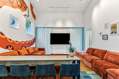 Bright modern living/media room with rust-orange velvet sectional sofas, wall-mounted TV, playful orange mural, teal chairs, potted plant and palm tree visible through windows.