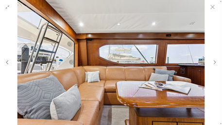 Sunlit luxury yacht salon with tan leather L-shaped sofa, textured gray pillows, glossy wood table with magazines, warm wood paneling and large windows framing a marina with boats.
