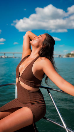 Smiling woman in a brown cutout dress basking in the sun on a boat railing, blue sky with fluffy clouds and a distant bridge over teal coastal water