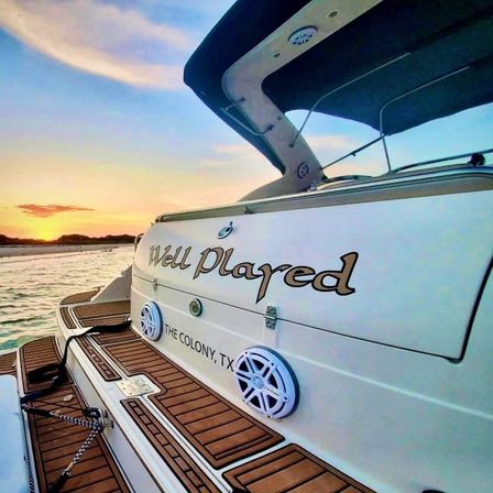 Stern of a white motorboat with teak swim platform, round marine speakers and painted name on the hull, anchored at sunset on a Texas lake near The Colony