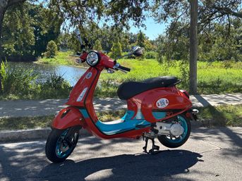 Red retro-style scooter with teal accents parked on a sunlit street beside a tree-lined pond and grassy park path.