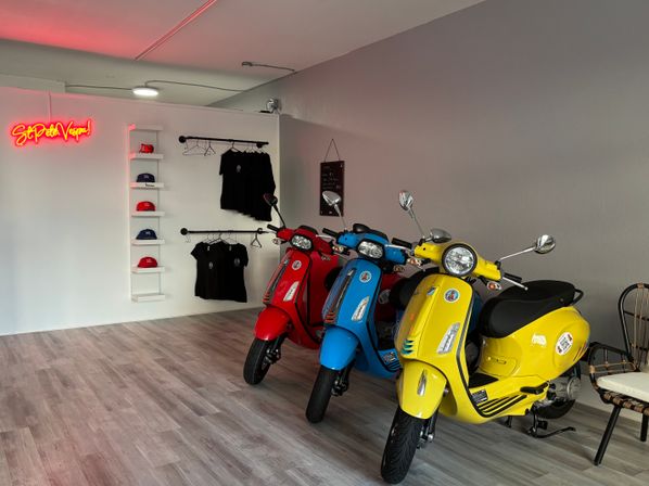 Three retro-style scooters—yellow, blue, and red—lined up in a bright modern showroom with wood-look flooring, a white display wall of hats and hanging black t-shirts, and a pink neon wall sign.