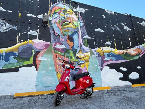 Red scooter parked in front of a vibrant, multicolored street mural of a serene figure with outstretched arms on a brick wall