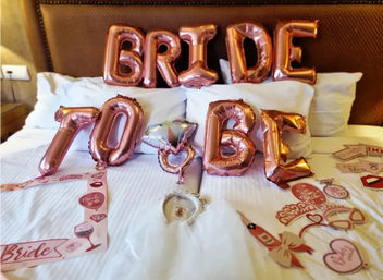 Rose-gold foil "BRIDE" and "TO BE" balloons on a hotel bed with tiara, bridal sash and bachelorette party props — festive bridal celebration setup.