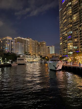 Waterfront marina at night with glowing high-rise condos, docked yachts, a lively boardwalk, and shimmering reflections on the water.