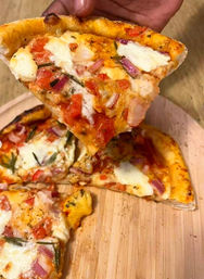 Hand lifting a slice of thin-crust pizza with melted mozzarella, diced tomatoes, red onion and rosemary, served on a round wooden pizza board.