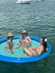 Three friends in bikinis relaxing in a blue inflatable floating pool on open water, wearing sun hats and sunglasses and toasting with champagne glasses, boats anchored in the background on a sunny summer day