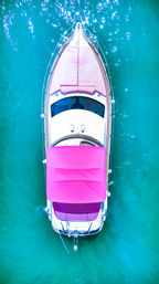 Aerial drone view of a white luxury yacht with a vibrant pink canopy floating on sparkling turquoise water, sunlit boating scene.
