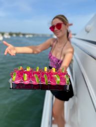 Tray of vibrant pink shot glasses garnished with lime wedges held aboard a boat, blurred marina and water in the background for a sunny summer party vibe