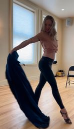 Woman striking a dramatic dance pose in a sunlit dance studio on hardwood floors, wearing a peach top, black leggings and strappy heels while swinging a flowing navy fabric.