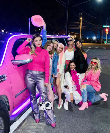 Eight friends in pink-themed outfits posing by a pink limousine with purple LED trim on a downtown street at night, one holding a cowboy hat and another wearing a feather boa and heart-shaped sunglasses.