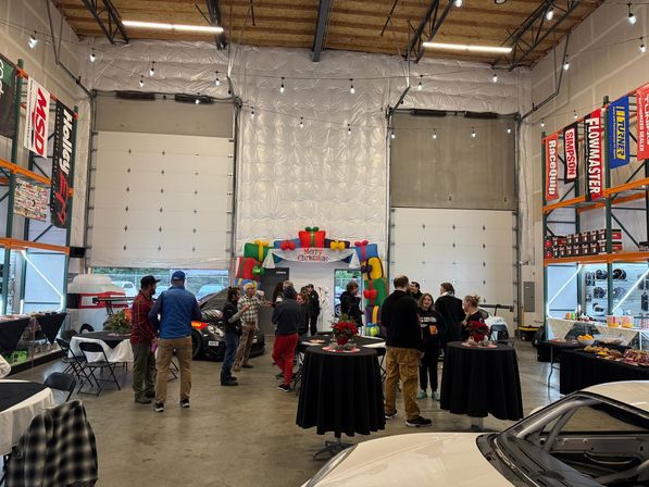 Auto shop holiday party in an open industrial garage—people mingling around high-top tables and a buffet, cars parked inside, a colorful 'Merry Christmas' inflatable arch, string lights, tall garage doors and automotive shelving with banners.