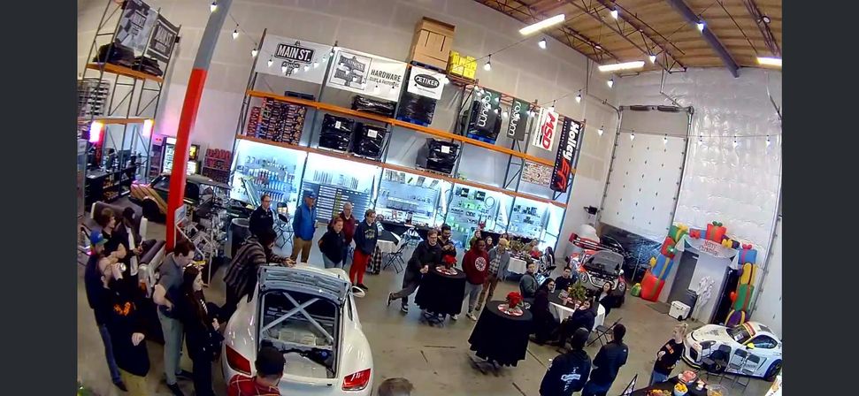 Warehouse-style motorsports garage open house with crowds mingling around two white race cars, high shelves and banners displaying auto parts, cocktail tables with snacks, and a colorful inflatable play area.